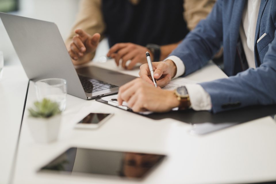 Two people at a bright desk working through figures together, one writing while the other reads from the laptop.