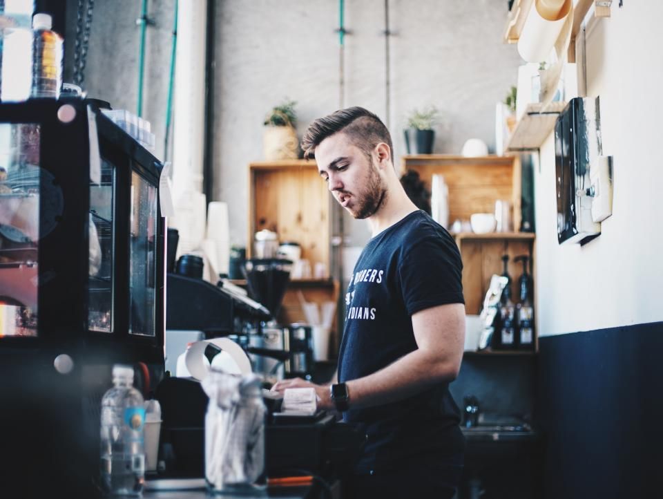 A barista at the espresso machine of his café, shelves of cups and supplies behind him.