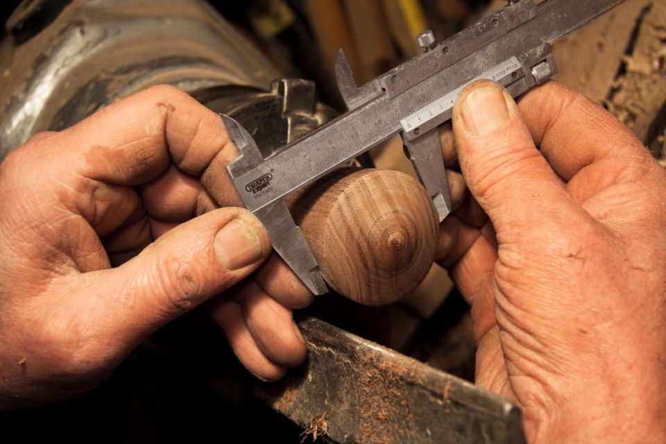 Two work-worn hands measuring a turned piece of wood with a pair of calipers.