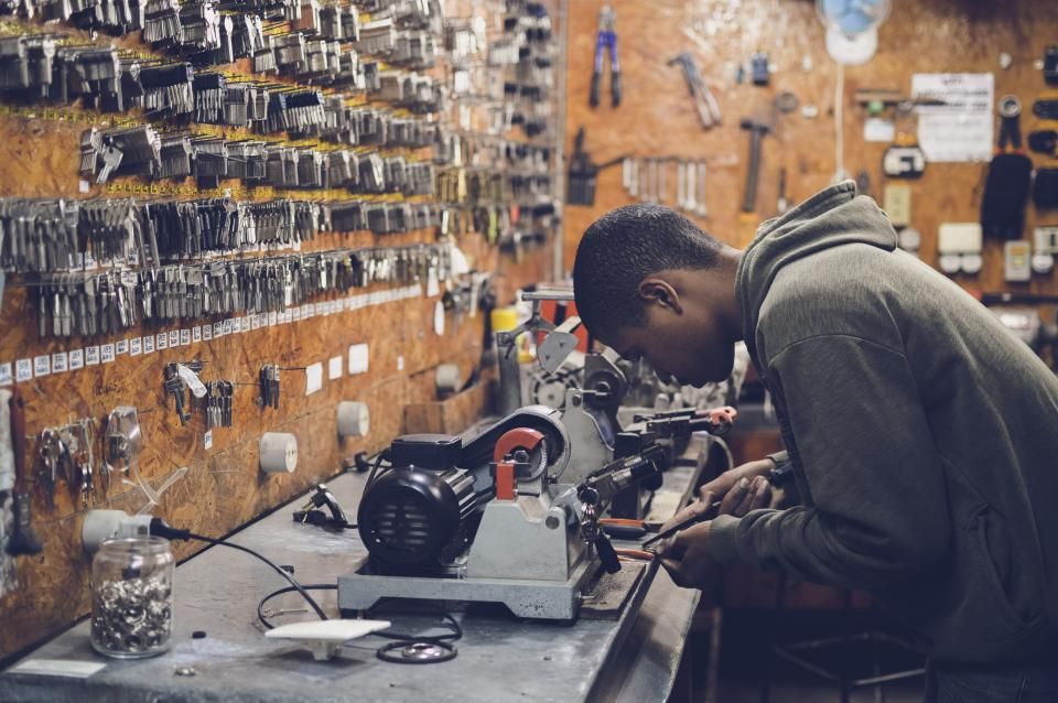 A young man working on a machine in a repair workshop, a wall of hanging tools behind him.