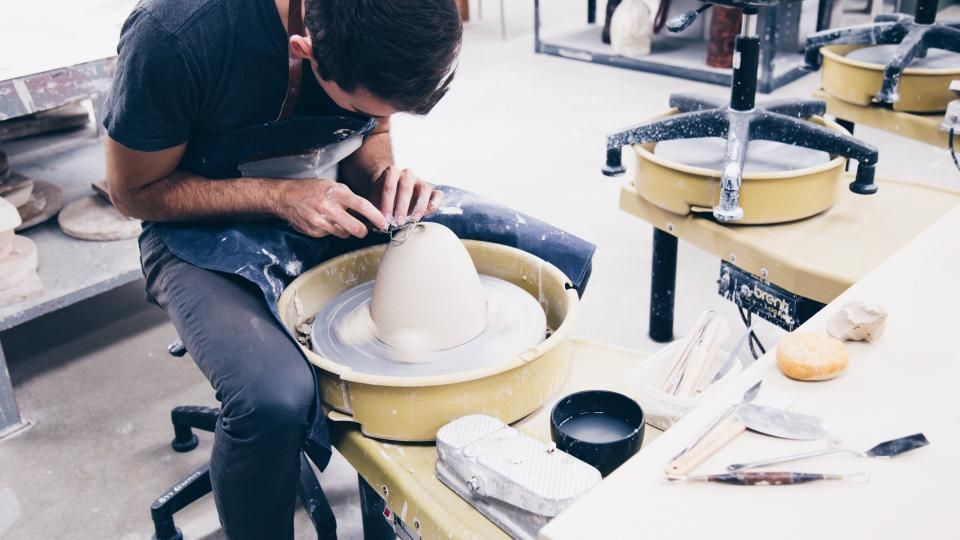 A maker sits at a potter's wheel in a bright workshop shaping a piece, with tools and other wheels around him.
